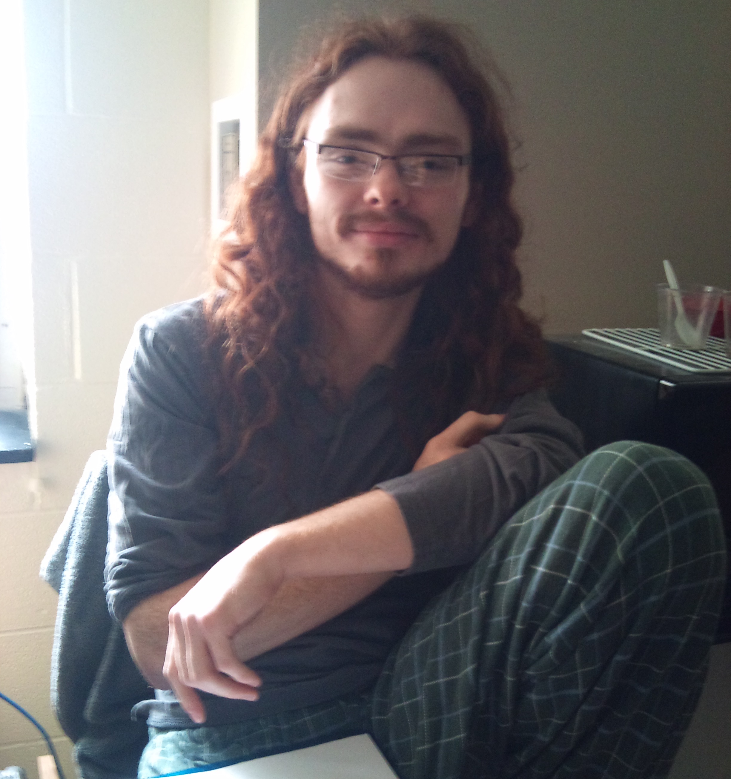 A photo of Eric smiling genuinely as he lounges between projects. He has long, curly red hair hanging loose, a scruffy goatee and mustache, and blocky, thin metal glasses.