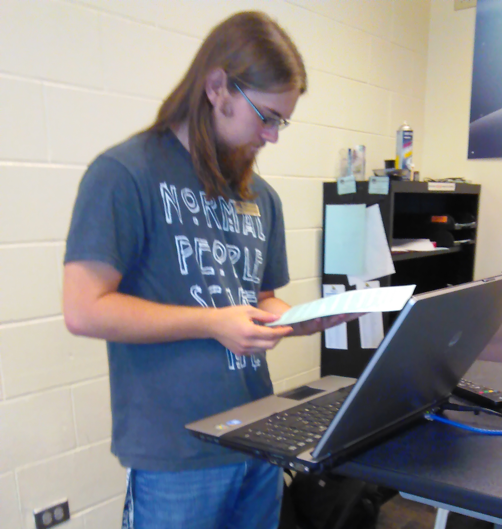 A photo of Nick concentrating on a work order with a laptop open in front of him. He has shoulder-length brown hair, thin glasses, and a thick goatee. His shirt reads, 'Normal People Scare Me.'