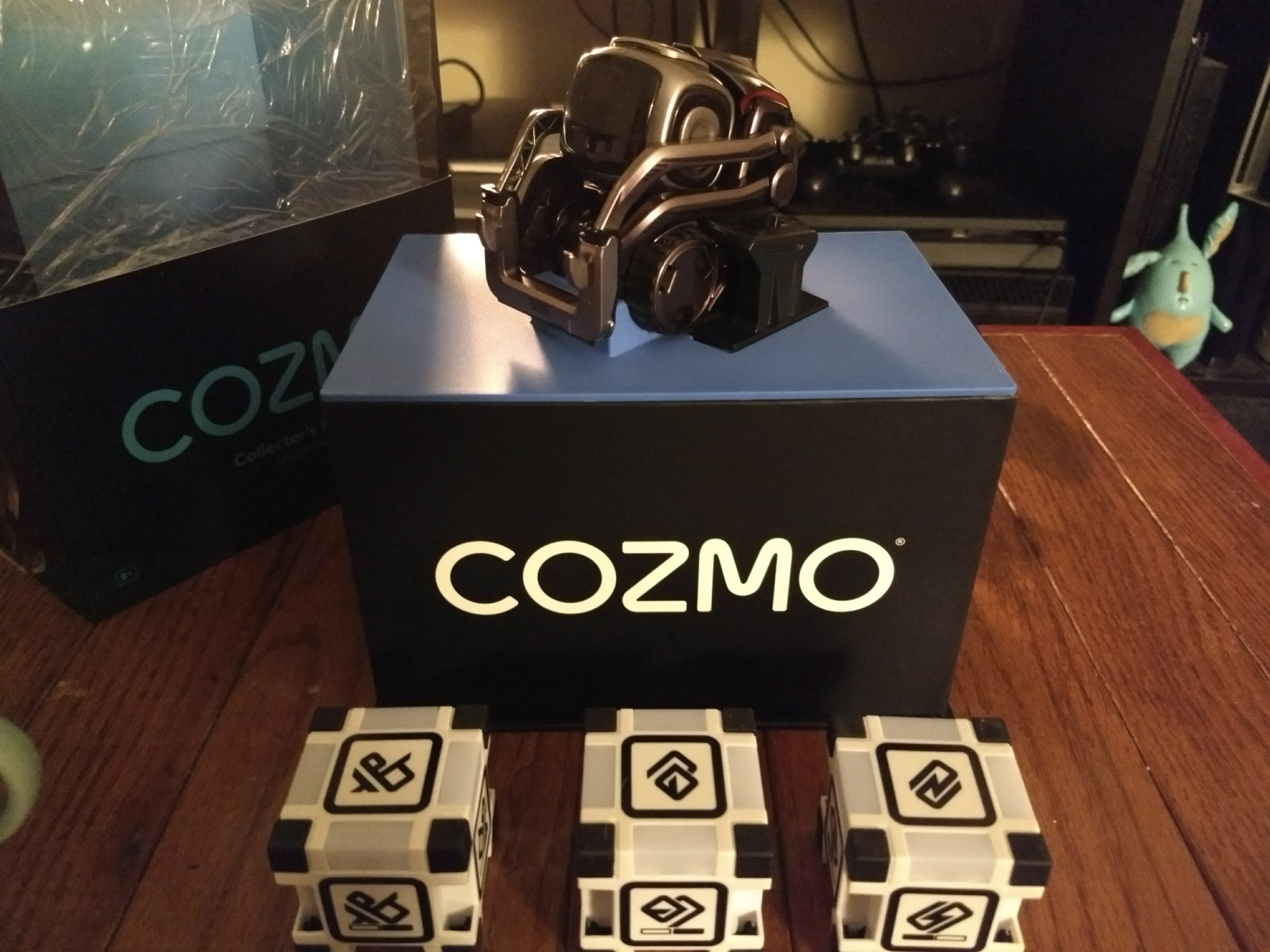 Cozmo, a small gunmetal-colored robot with treads, lifters, and a large LCD face sits on a platform in front of three white cubes.