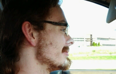 A photo of Eric. He has long, curly red hair, a scruffy goatee and mustache, and blocky, thin metal glasses.